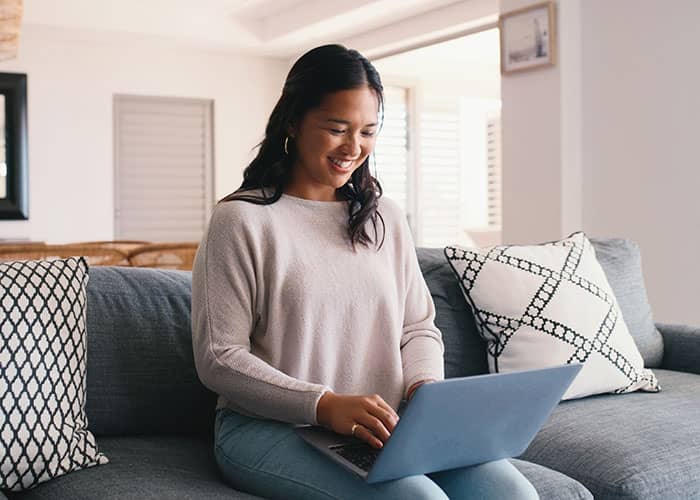 A smiling prospective student sitting on a sofa with a laptop, researching degrees and applying to Rhodes State College online.